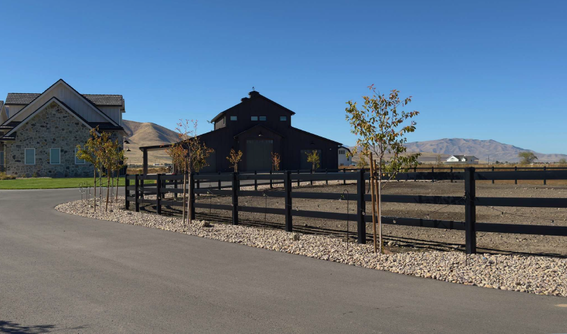 Rural road beside black ranch buildings and a wooden fence under a clear blue sky.