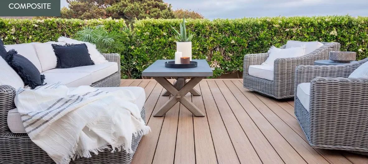 A gray wicker outdoor sofa and two armchairs with white cushions on a wooden deck with a central coffee table and greenery.