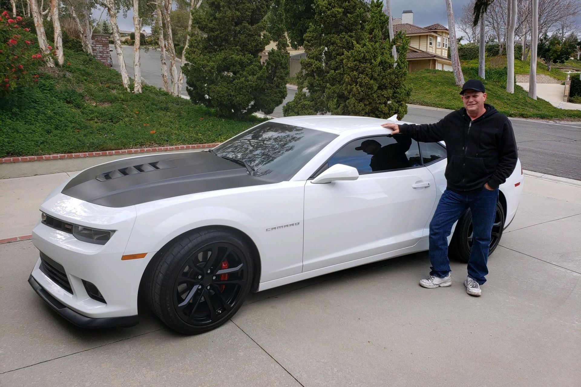 Man leaning on a white sports car with black hood and wheels on a suburban driveway