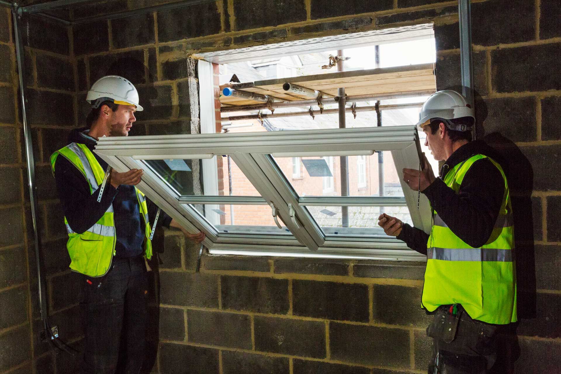 Two workers in safety vests and hard hats installing a skylight in a brick wall opening