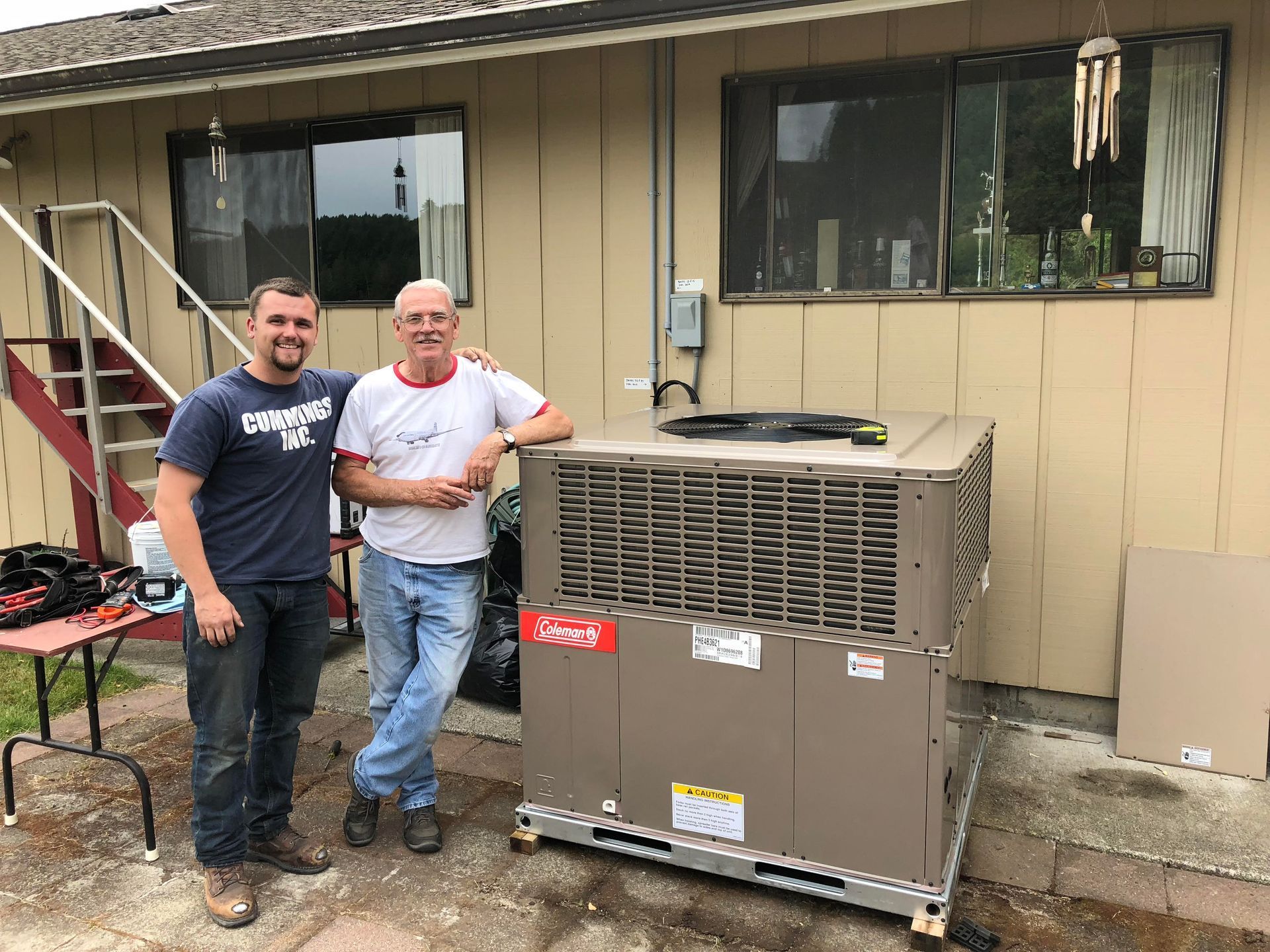 Two men pose with a new air conditioning unit outside a building with two windows.
