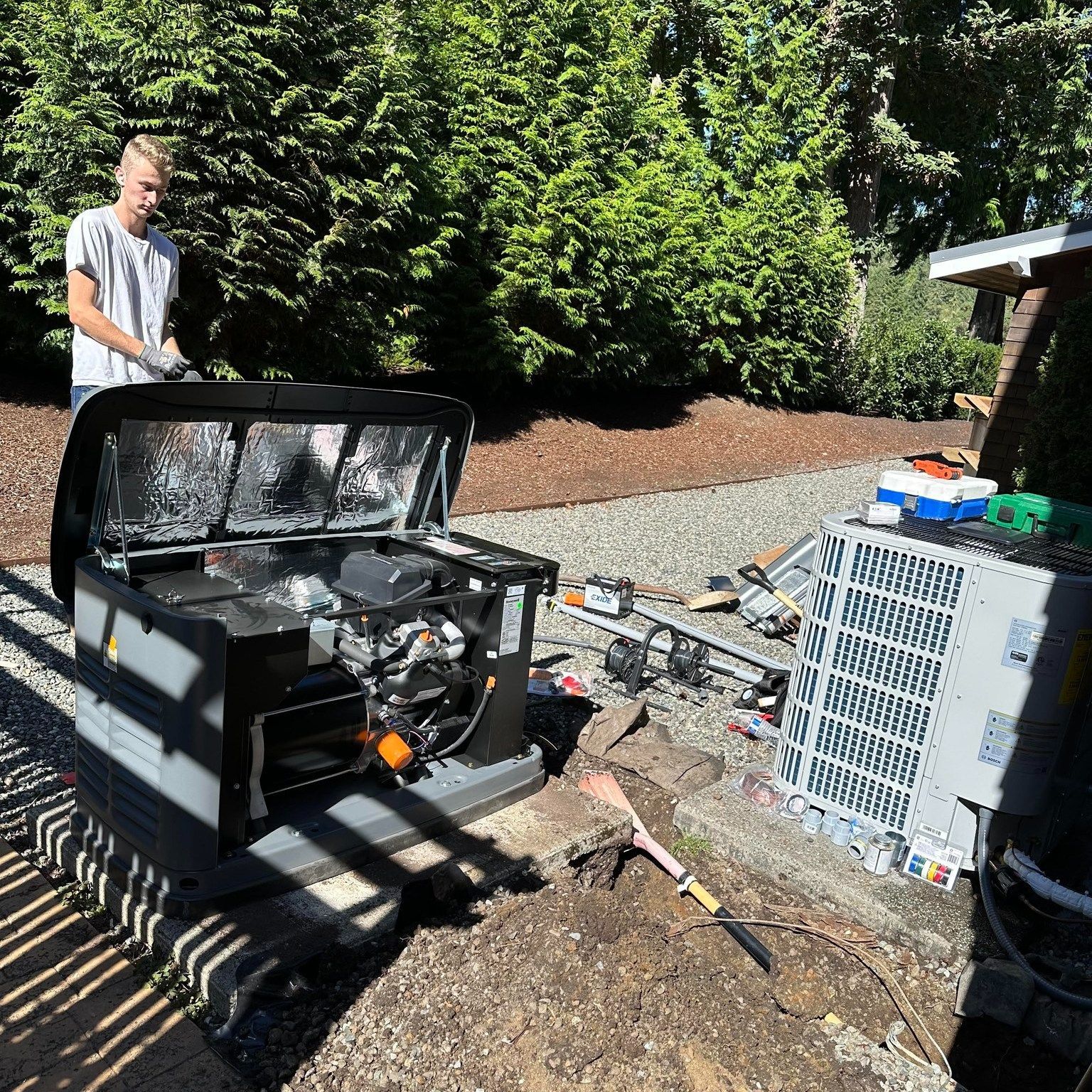 Man standing near open generator in a backyard. Various equipment and tools scattered around.