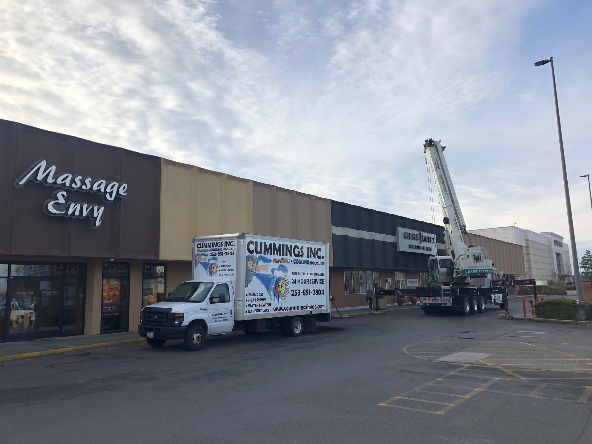 A crane lifts a sign from a Massage Envy store next to a truck. Cloudy sky, commercial setting.