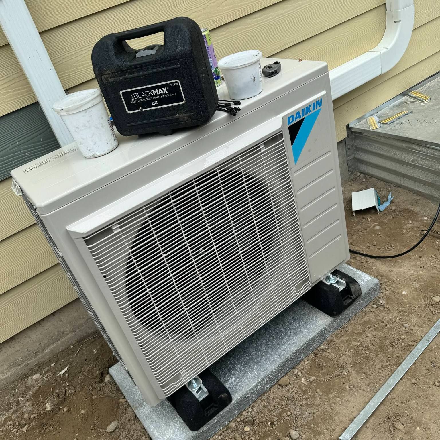 Daikin heat pump unit on a concrete block, next to a beige building, with tools and a gutter.