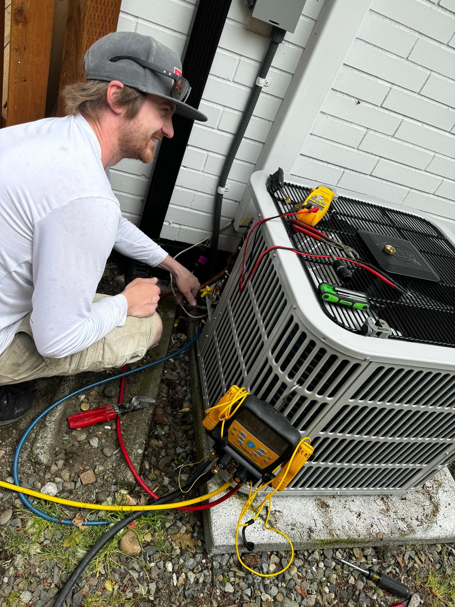 HVAC technician repairs an air conditioner unit outside. He connects gauges and electrical lines while smiling.