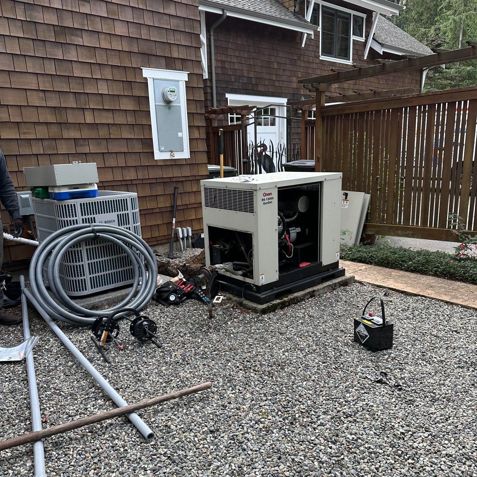 Generator installation in a gravel yard, next to a house with brown siding. Electrical components are visible.