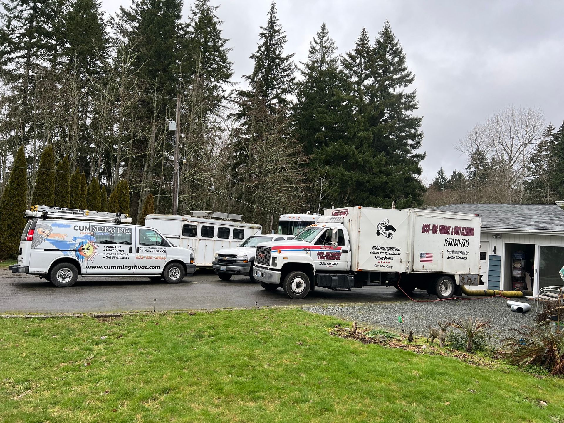 Vehicles parked near a building: a service truck, a van, and trailers, in front of trees.