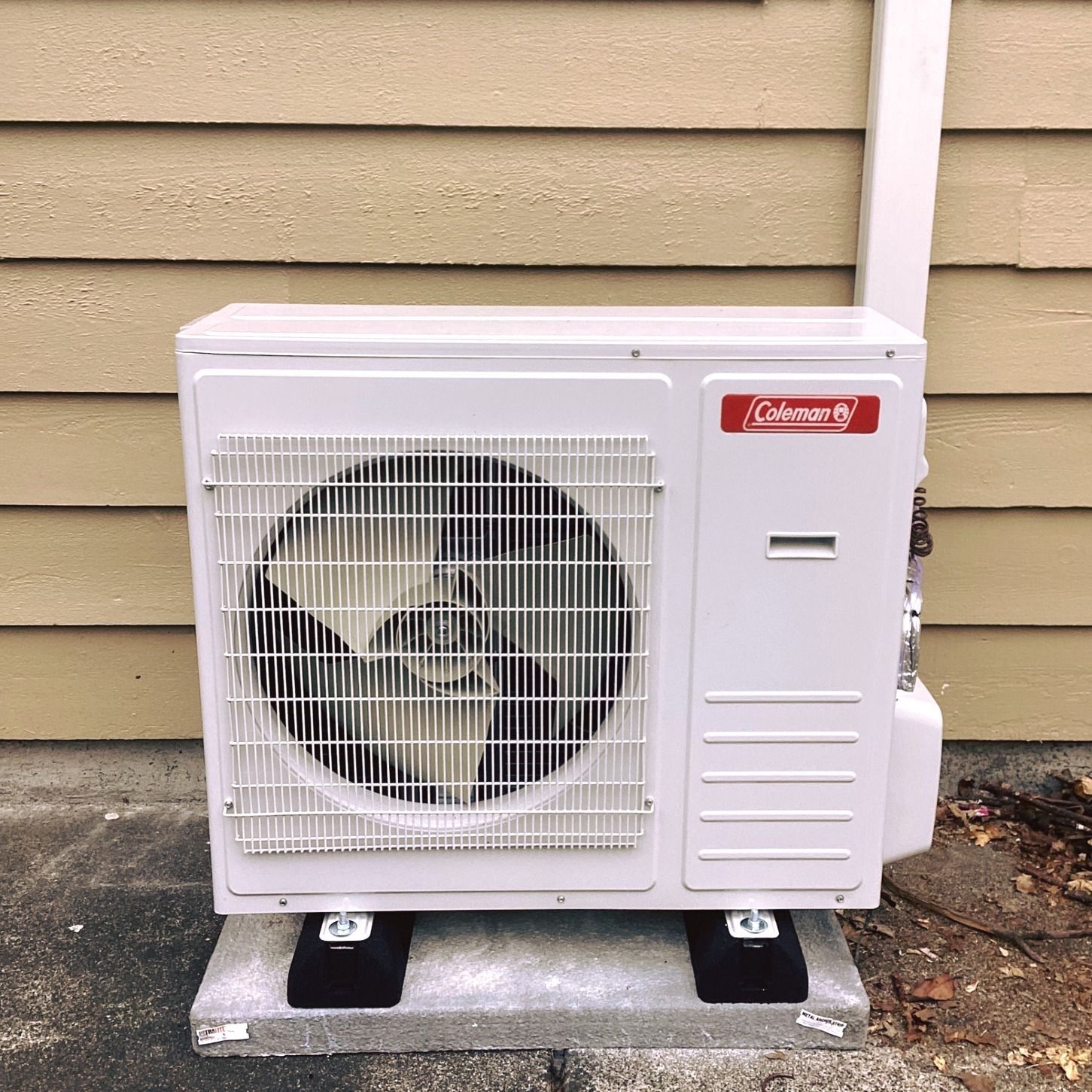 White Coleman air conditioner unit mounted on black supports against a beige siding wall.