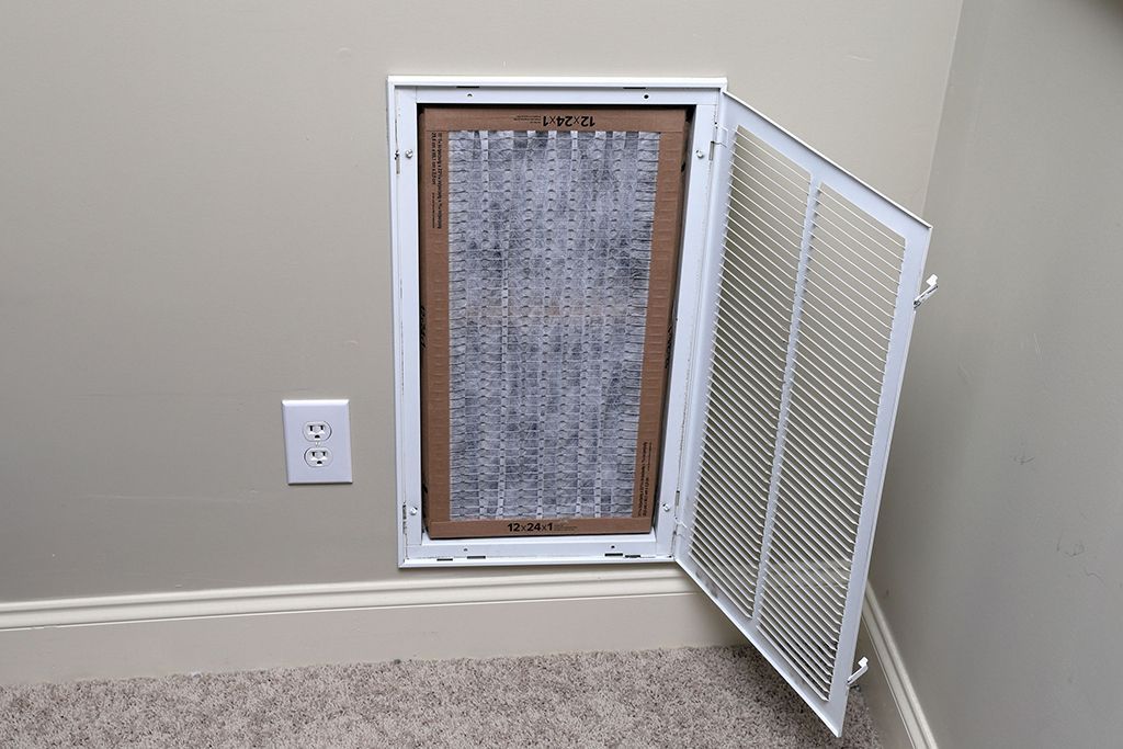 HVAC air filter, dirty and inside a white cabinet on a beige wall with a partially open metal grate.