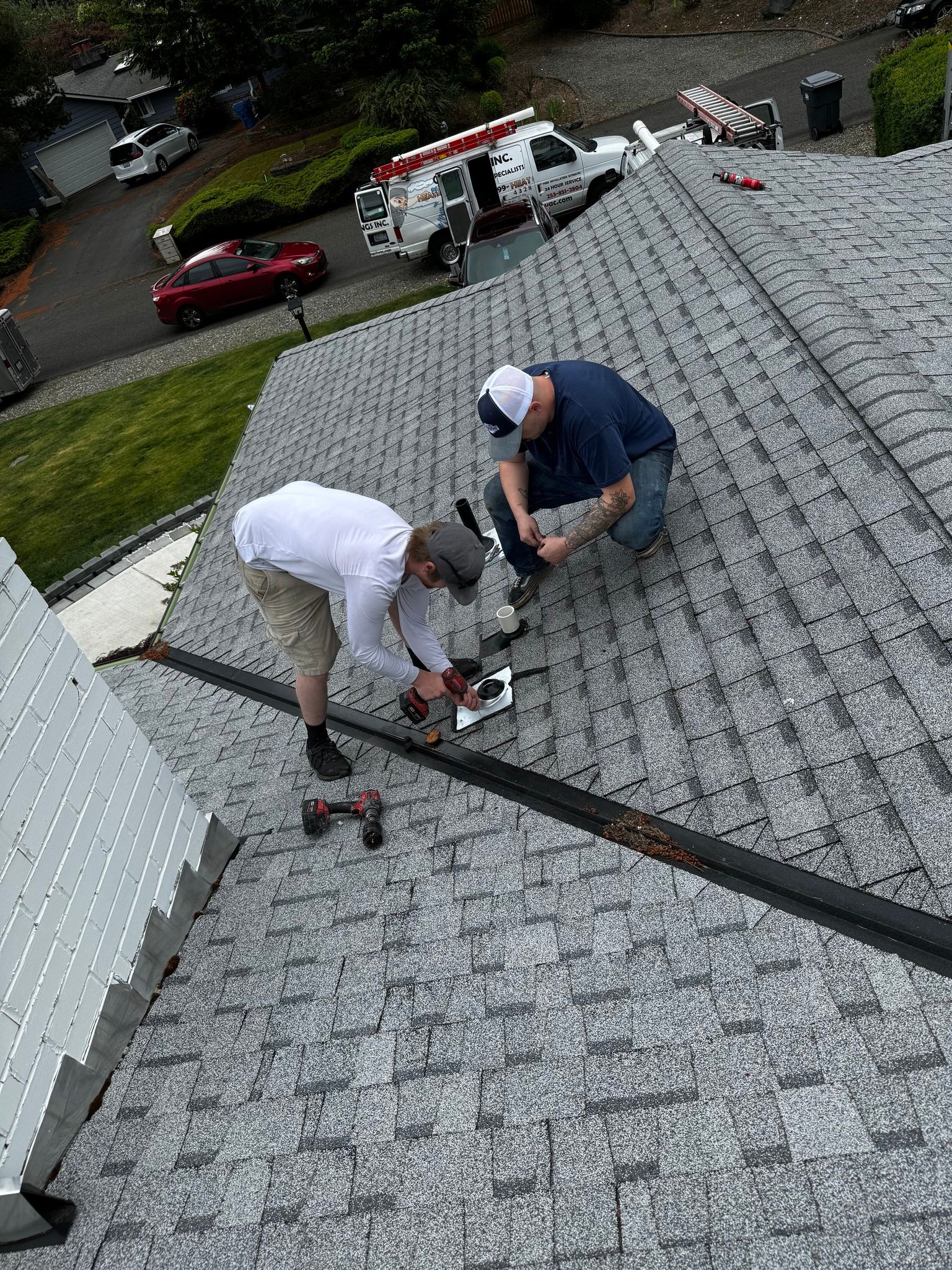 Two people on a roof working, with a truck and cars in the background.