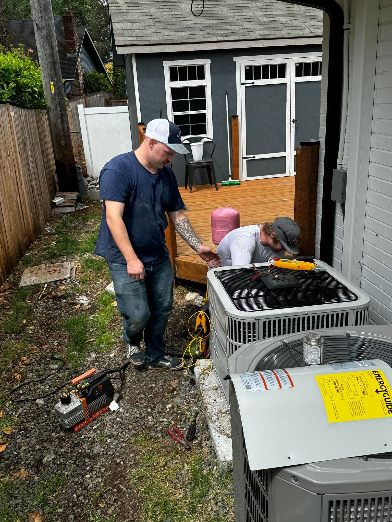 Two men working on HVAC units outdoors near a gray building. One points at the unit.