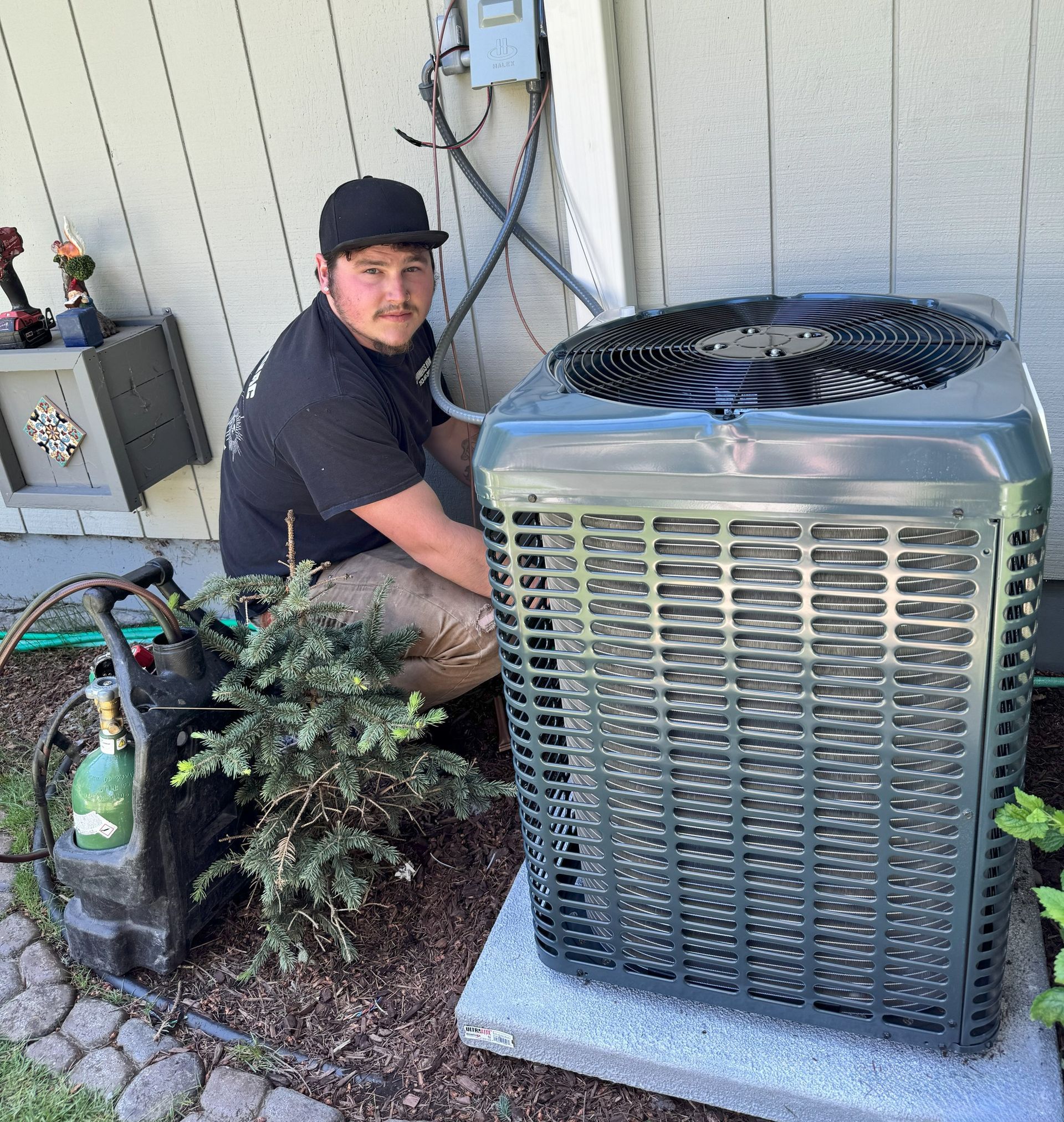 HVAC technician kneels near an AC unit, working outdoors. He wears a black cap and shirt.