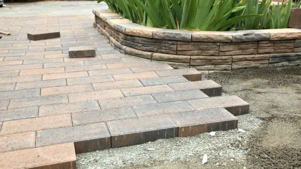 Brick walkway under construction next to a curved stone planter with green plants.