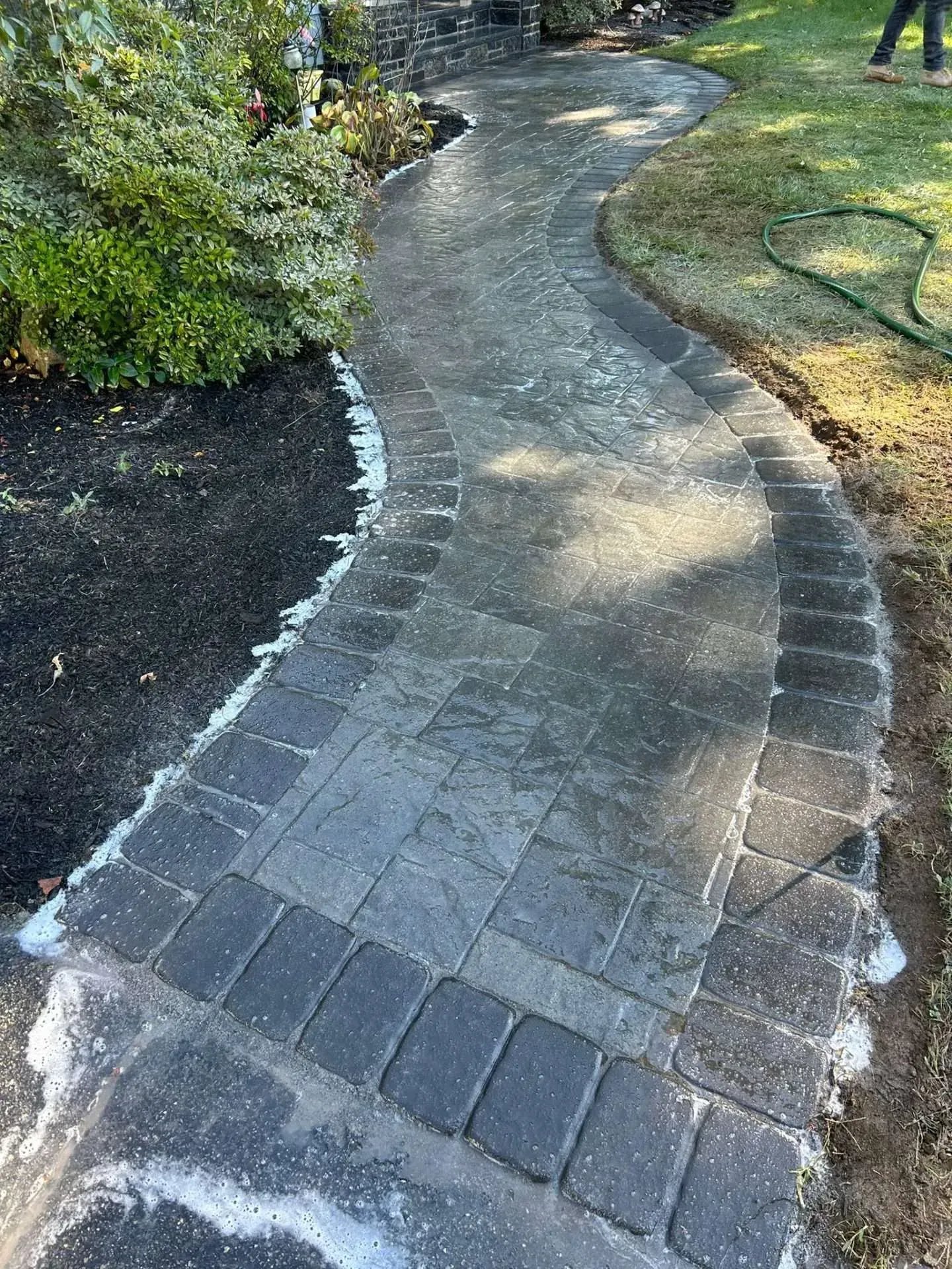 A winding brick pathway with dark border, wet from cleaning, alongside garden and lawn.