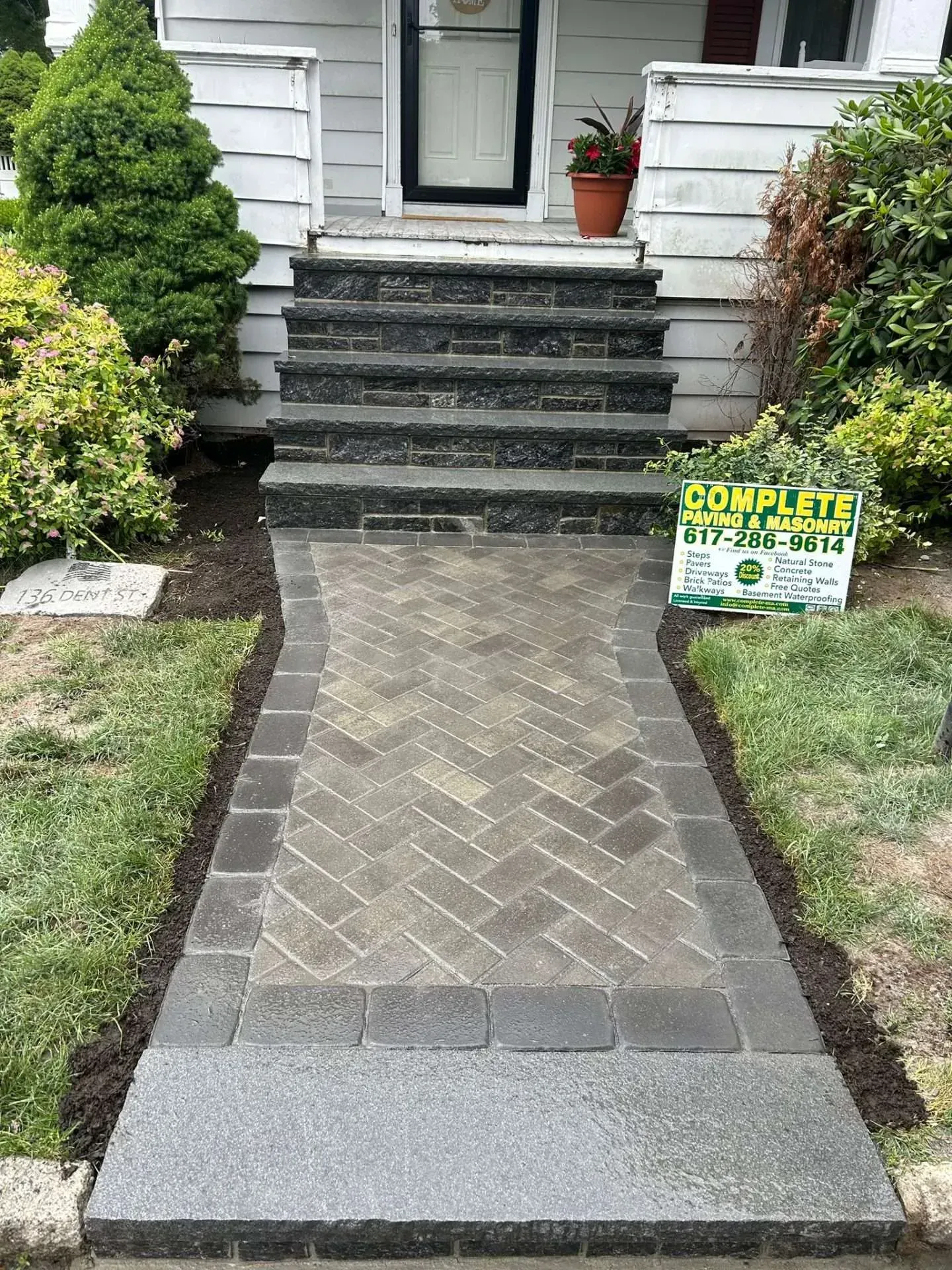 Brick pathway leading to steps, front door. Dark gray bricks, green grass, bushes. 