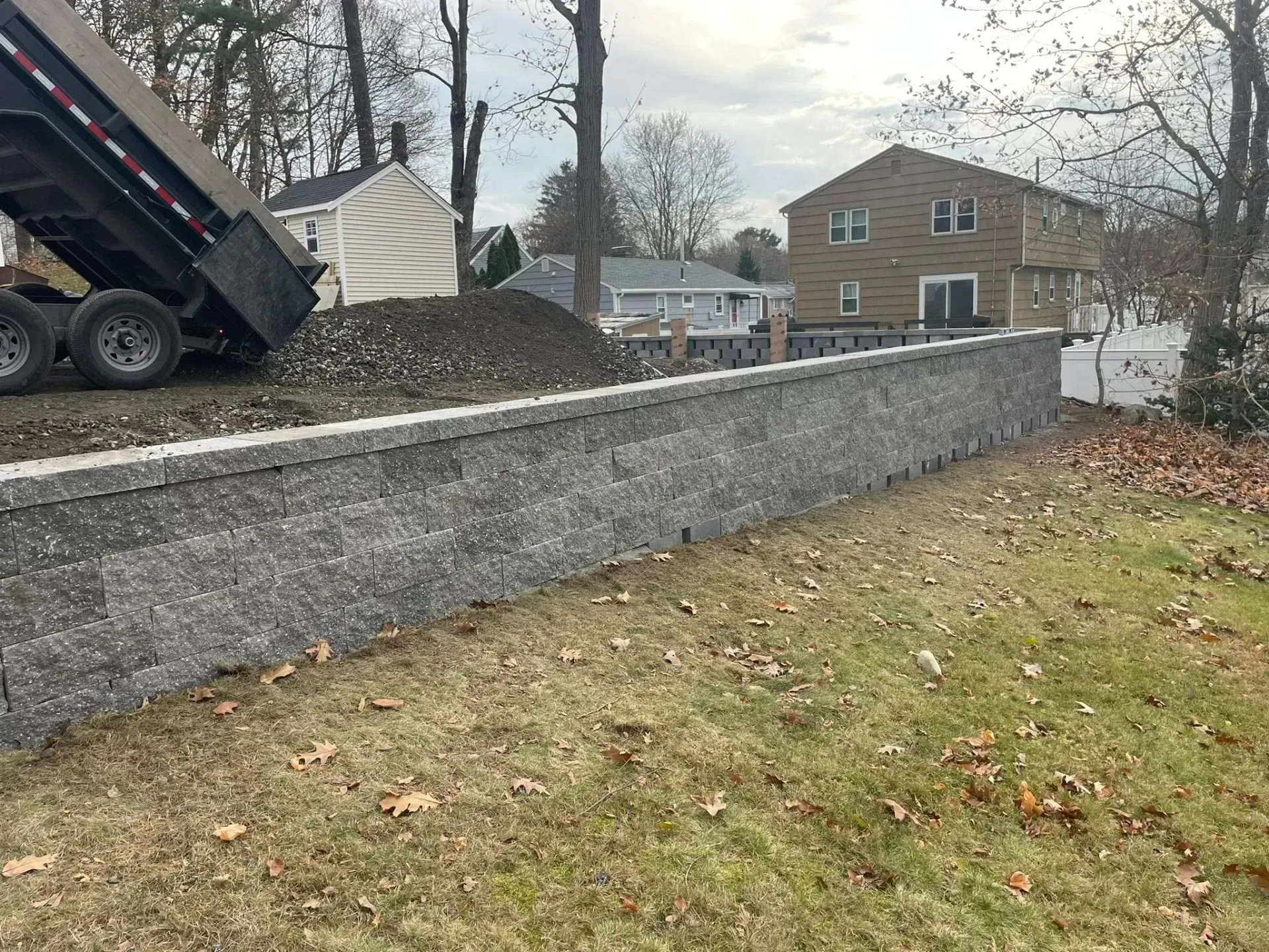 Gray stone retaining wall with a truck dumping gravel; houses in background.