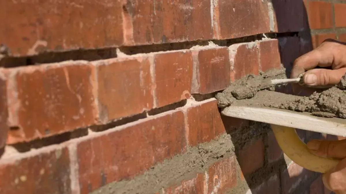 A person uses a trowel to apply mortar to the joints of a red brick wall.