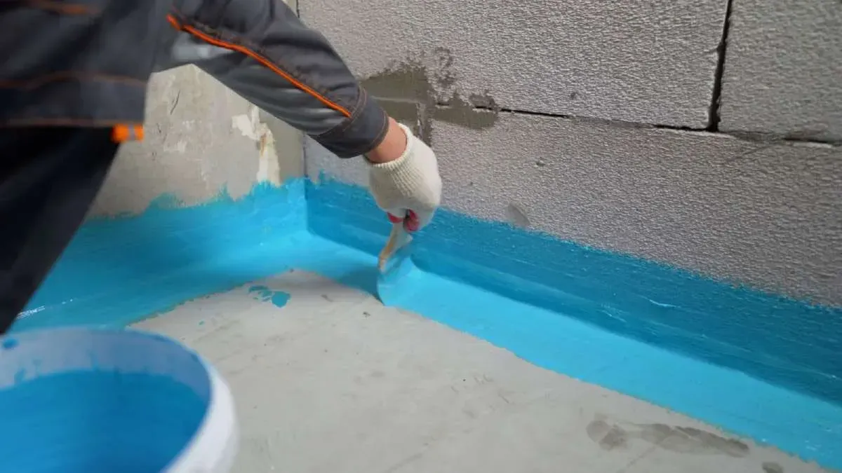 Person applying blue sealant to a wall corner. Bucket of sealant and trowel present. Gray concrete block walls.