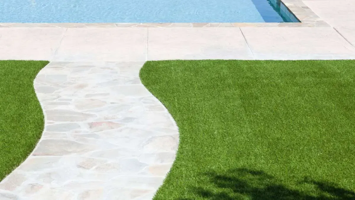 Stone pathway curves through green grass next to a light-colored pool deck and blue water.