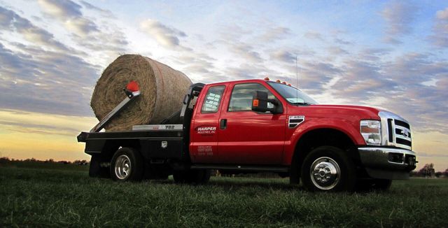 Red flatbed truck hauling a large hay bale across a grassy field at sunset