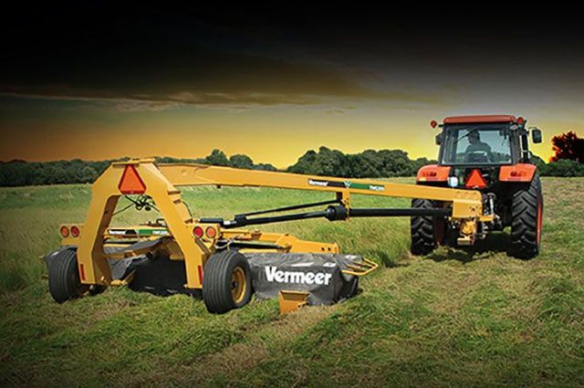 Yellow Vermeer mower attachment behind an orange tractor in a grassy field under a cloudy sky