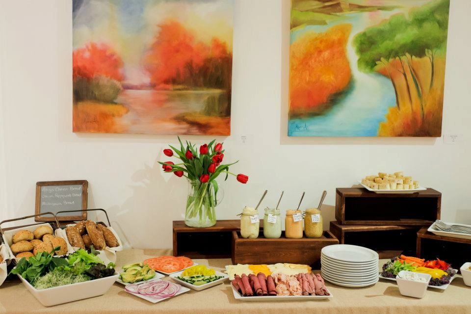 Buffet table with food, flowers, and artwork. A variety of dishes are arranged with colorful paintings hanging above.