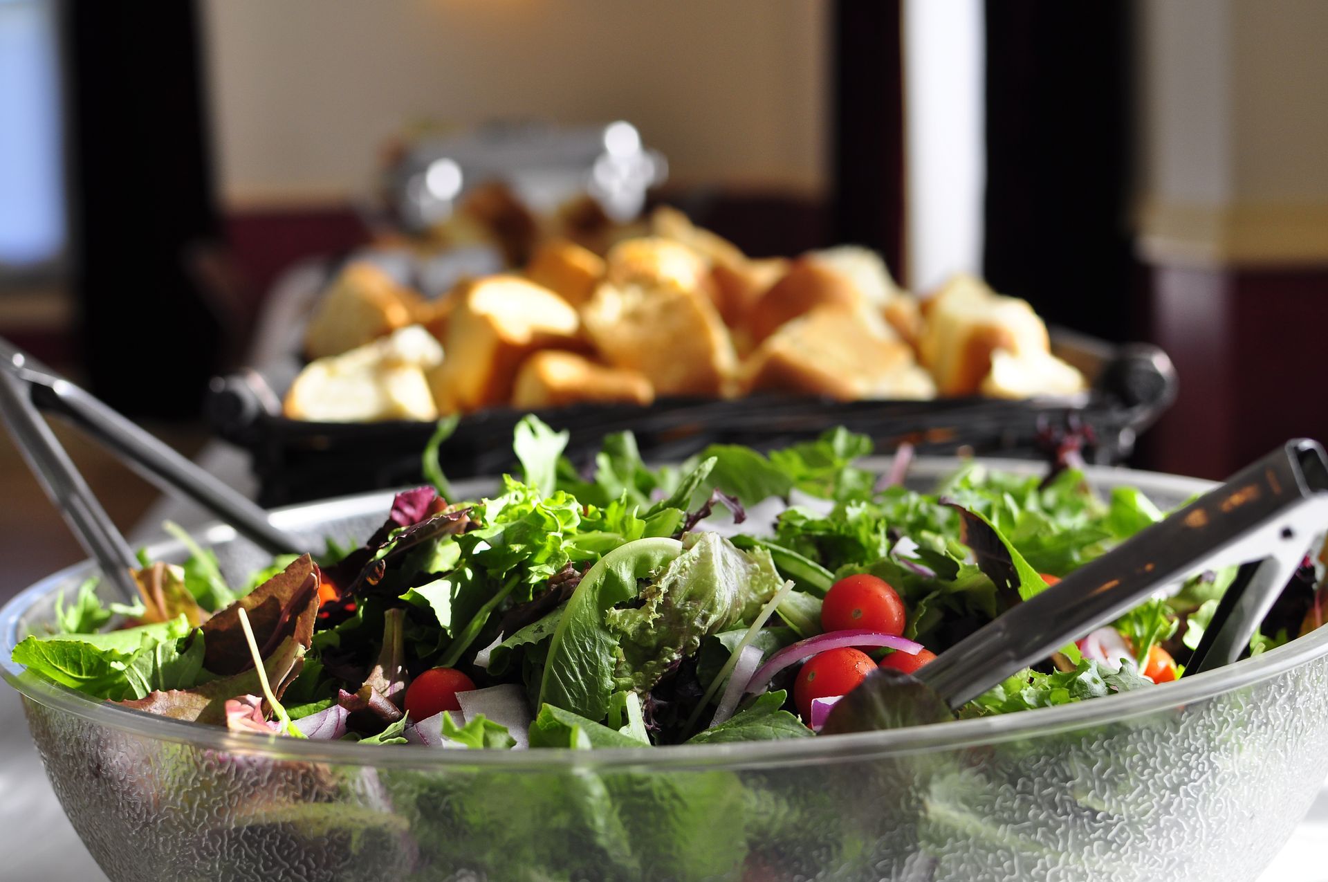 Large salad bowl with greens, tomatoes, red onion, and bread basket in background.