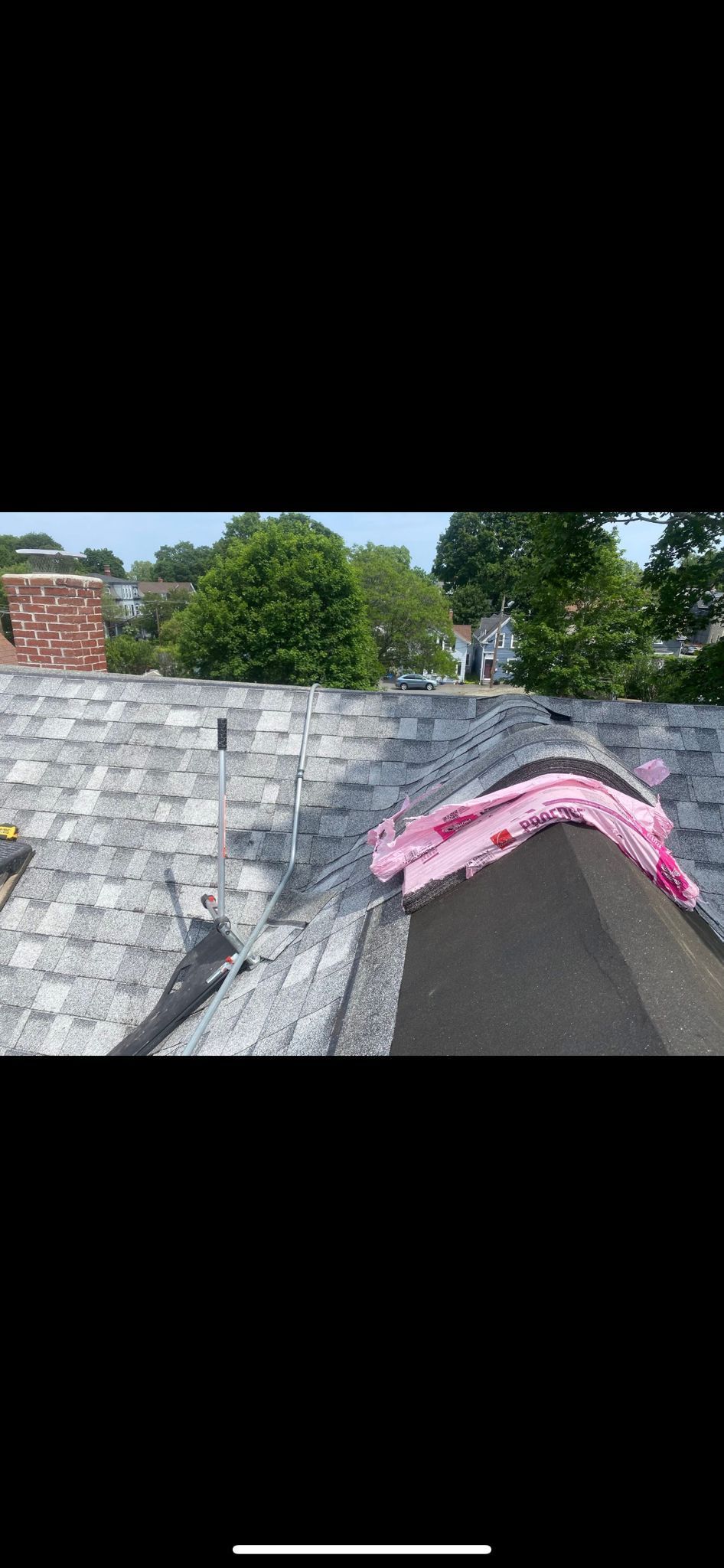 A roof with gray shingles and pink foam at the edge. Trees and a brick building are in the background.