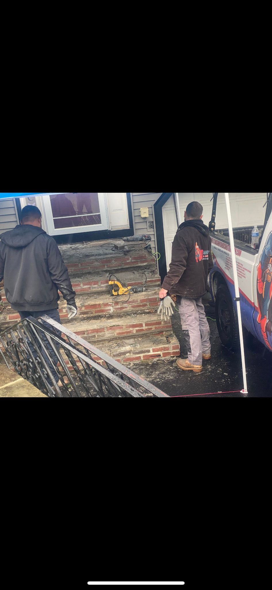 Two men walking on a debris-filled ground near a white vehicle. One man wears a dark jacket, the other a brown one.
