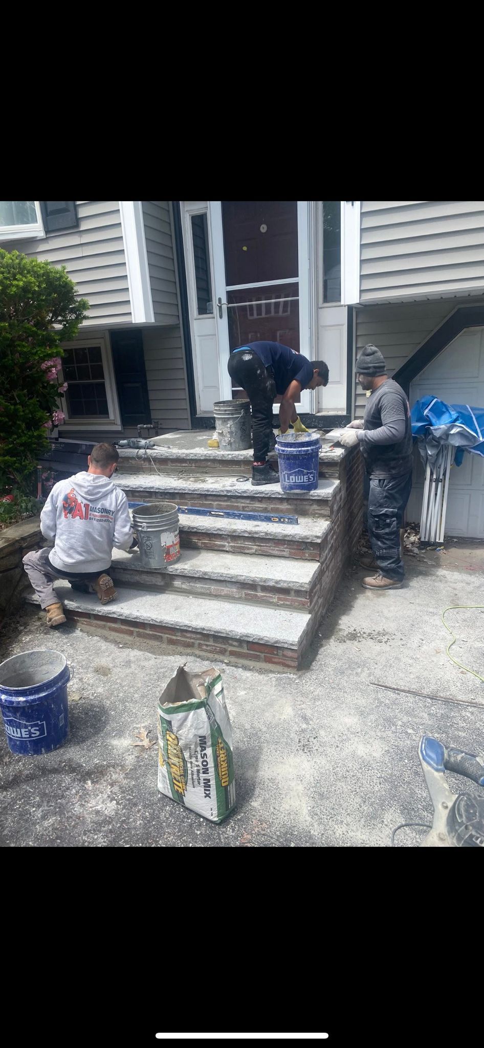 Workers laying stone steps at a house entrance. They are mixing cement, using buckets, and on their knees.