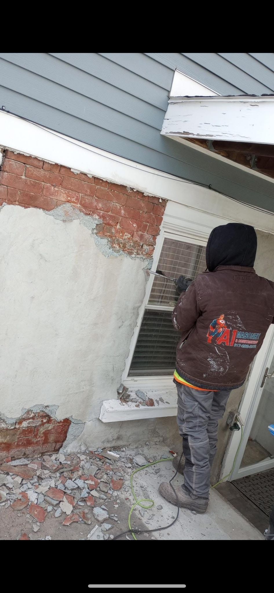 A person in work clothes near a building with exposed brick, stucco, and a window.