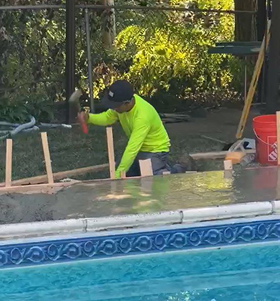 A construction worker spreads concrete next to a pool, wearing a neon green shirt and black cap.