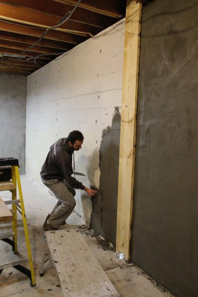 Man applying gray plaster to wall in a basement. He is using a trowel, standing on a plank.