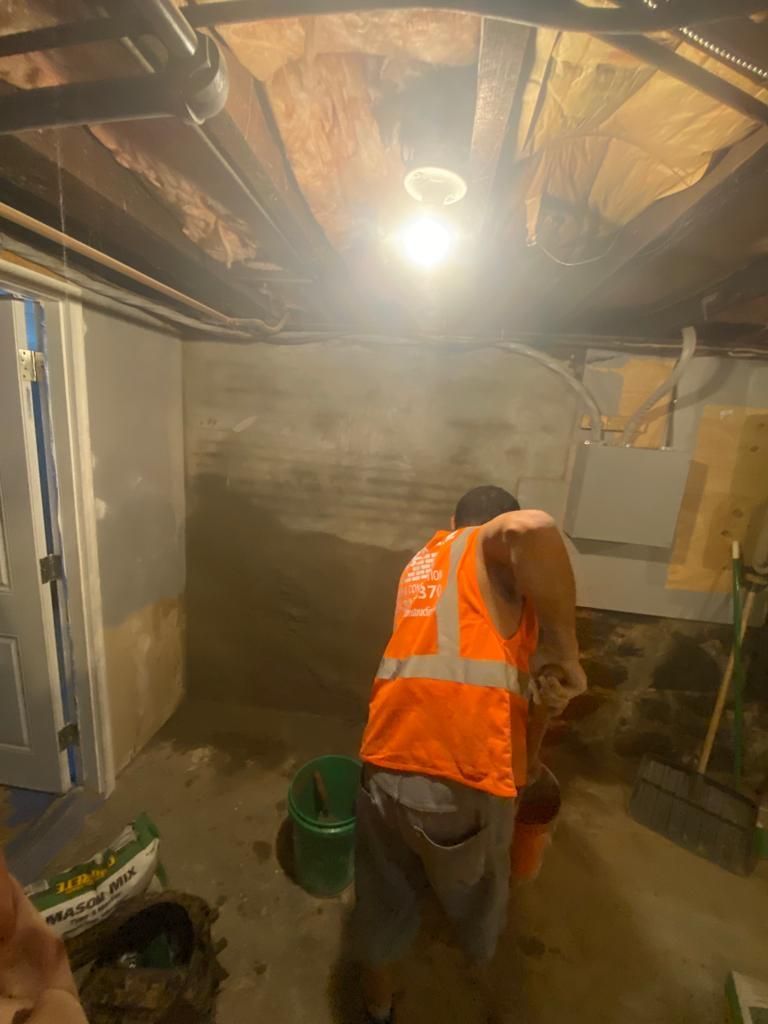 Man in orange vest working on basement wall. Dark, dimly lit space.