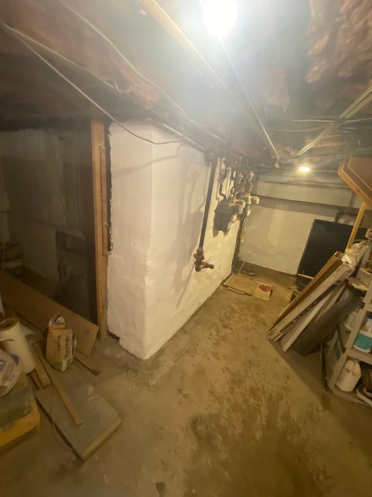 Basement interior with white walls, exposed pipes, and wooden beams. Construction materials scattered around.