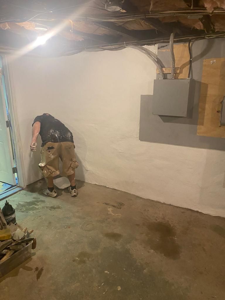 Person painting a basement wall; gray cement floor; utility box on the right; dim lighting.