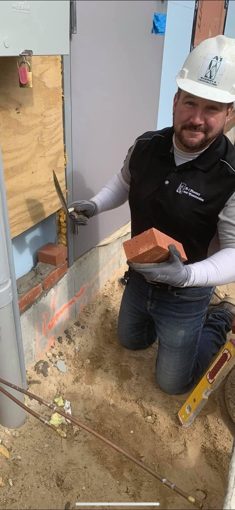 A bricklayer, kneeling, holds a brick. He wears a hard hat, gloves, and a work vest. Construction site.