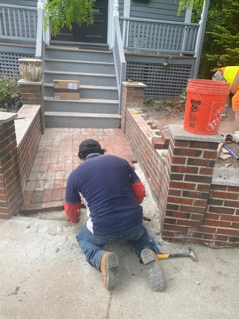 Man laying brick path in front of a house. Red brick walls, concrete stairs and path, orange bucket visible.