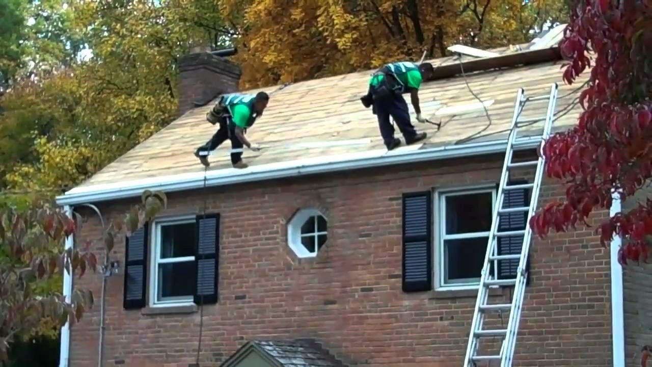 Two roofers replacing shingles on a brick house roof with fall foliage visible.