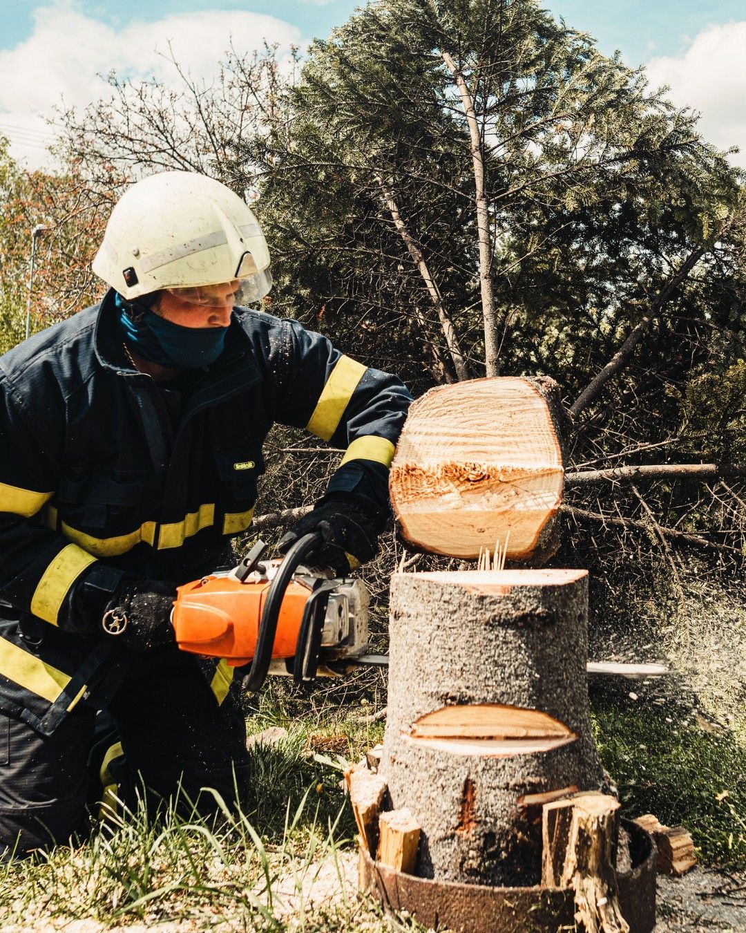 Firefighter in uniform uses a chainsaw to cut a log. He wears a helmet and is outdoors, near a tree.