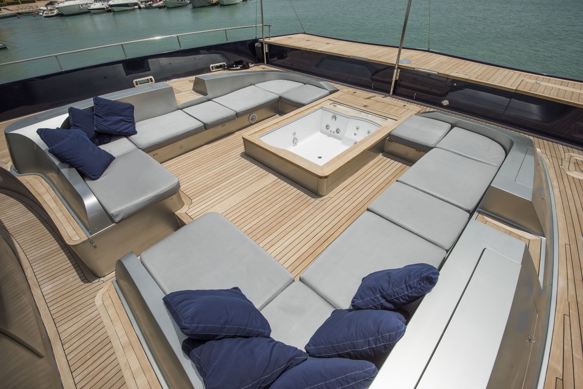 Yacht deck with gray lounge seating around a hot tub, blue pillows. Wooden deck, sunny day.