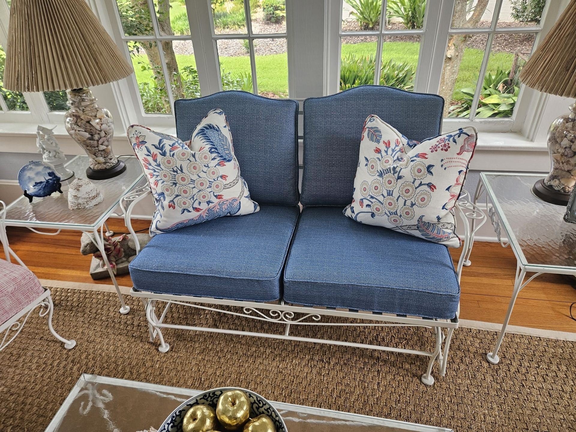 A blue loveseat with floral pillows sits between glass-topped tables in a sunroom with a view of a garden.