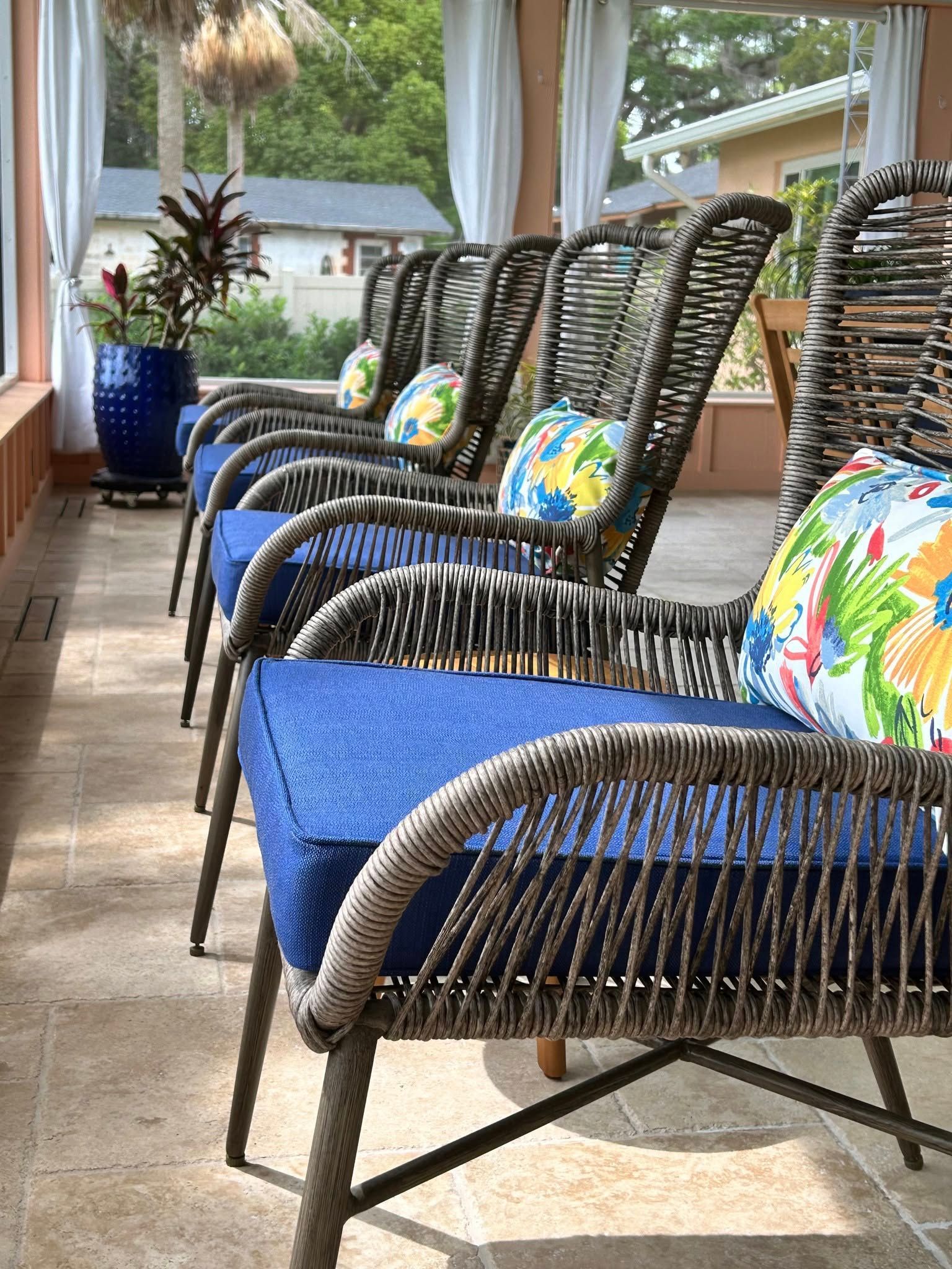 Row of woven chairs with blue cushions, colorful pillows, and a blue planter on a porch.