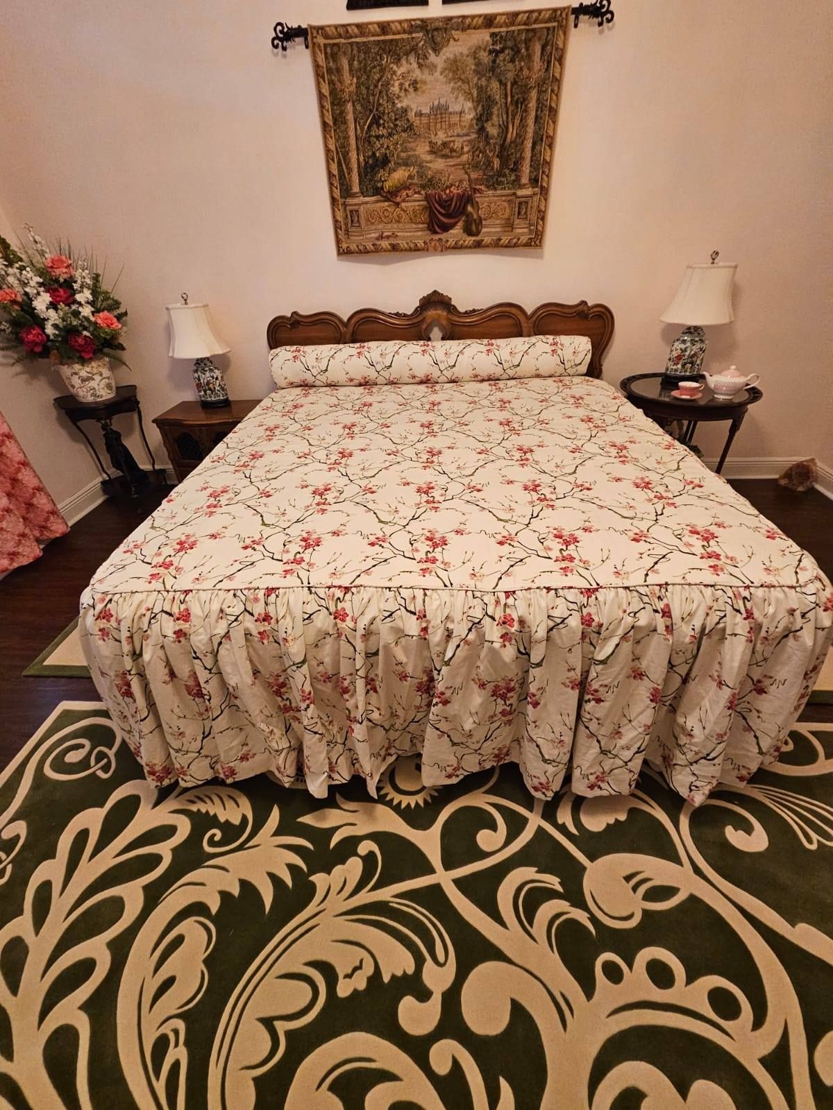 Bedroom with floral bedspread, ornate headboard, lamps, tapestry, and patterned green rug.