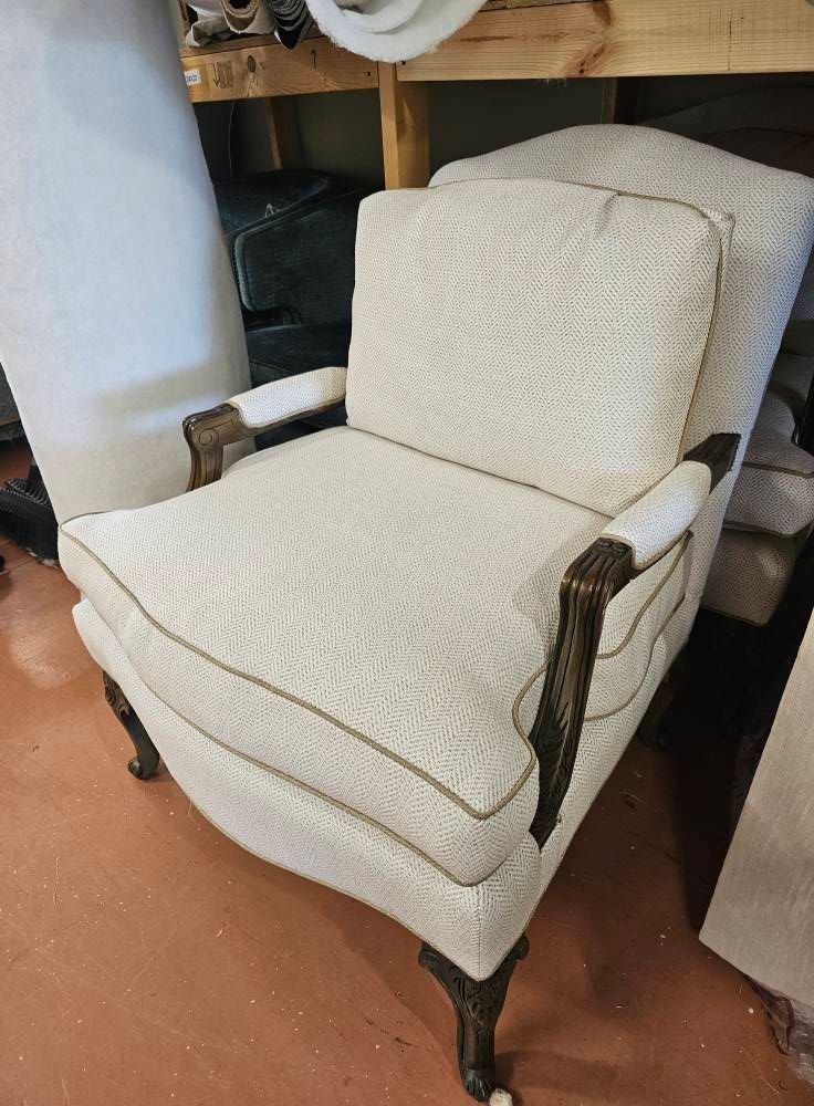 Cream-colored armchair with patterned fabric, dark wood arms and legs, gold trim, in a workshop.