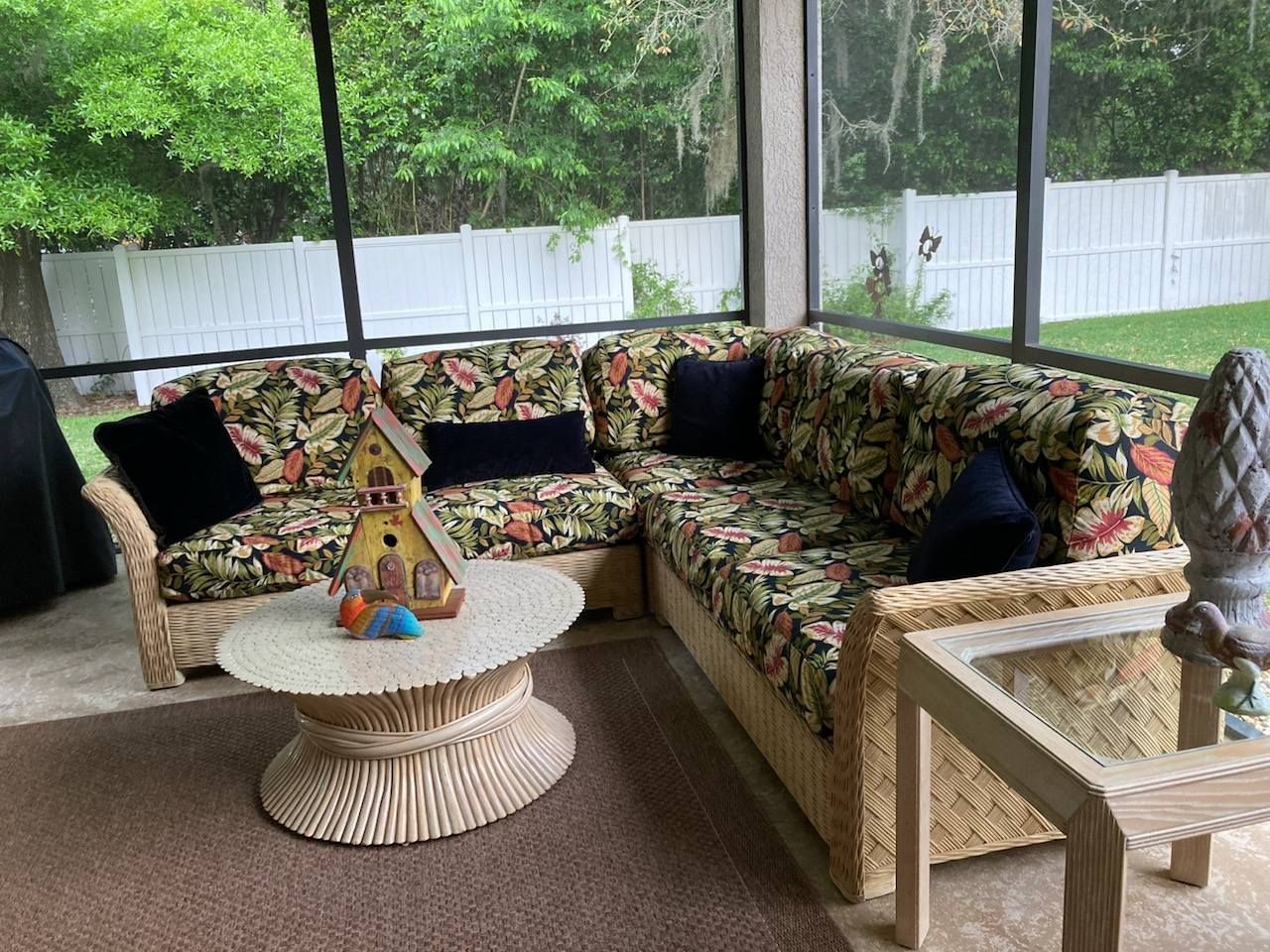 L-shaped wicker sofa with floral cushions and black pillows on a screened porch with a round table and yard.