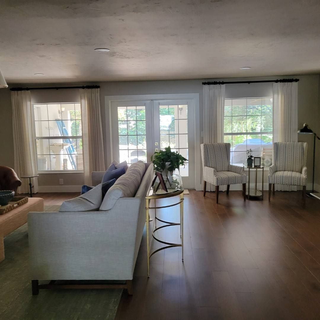 Living room with light-colored furniture, hardwood floors, and large windows with white curtains.