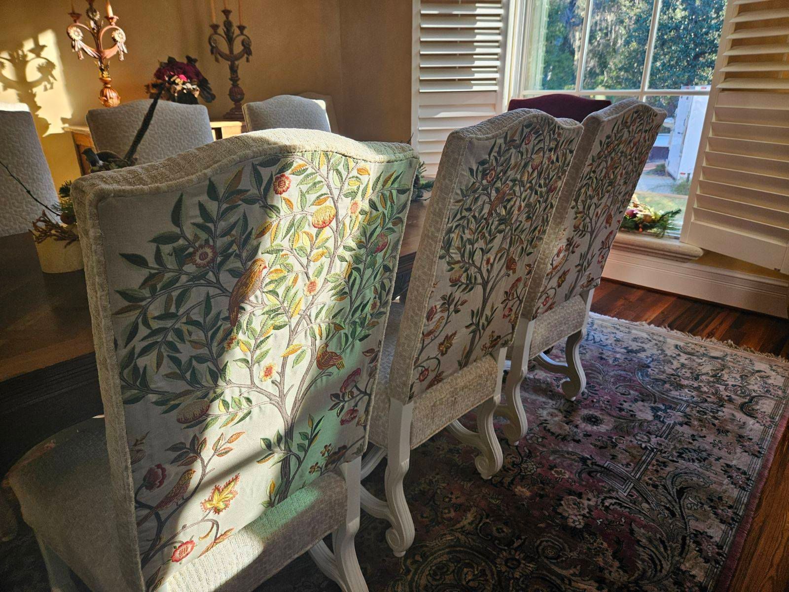 Dining chairs with floral upholstery are positioned around a table, a rug beneath. Sunlight streams in.