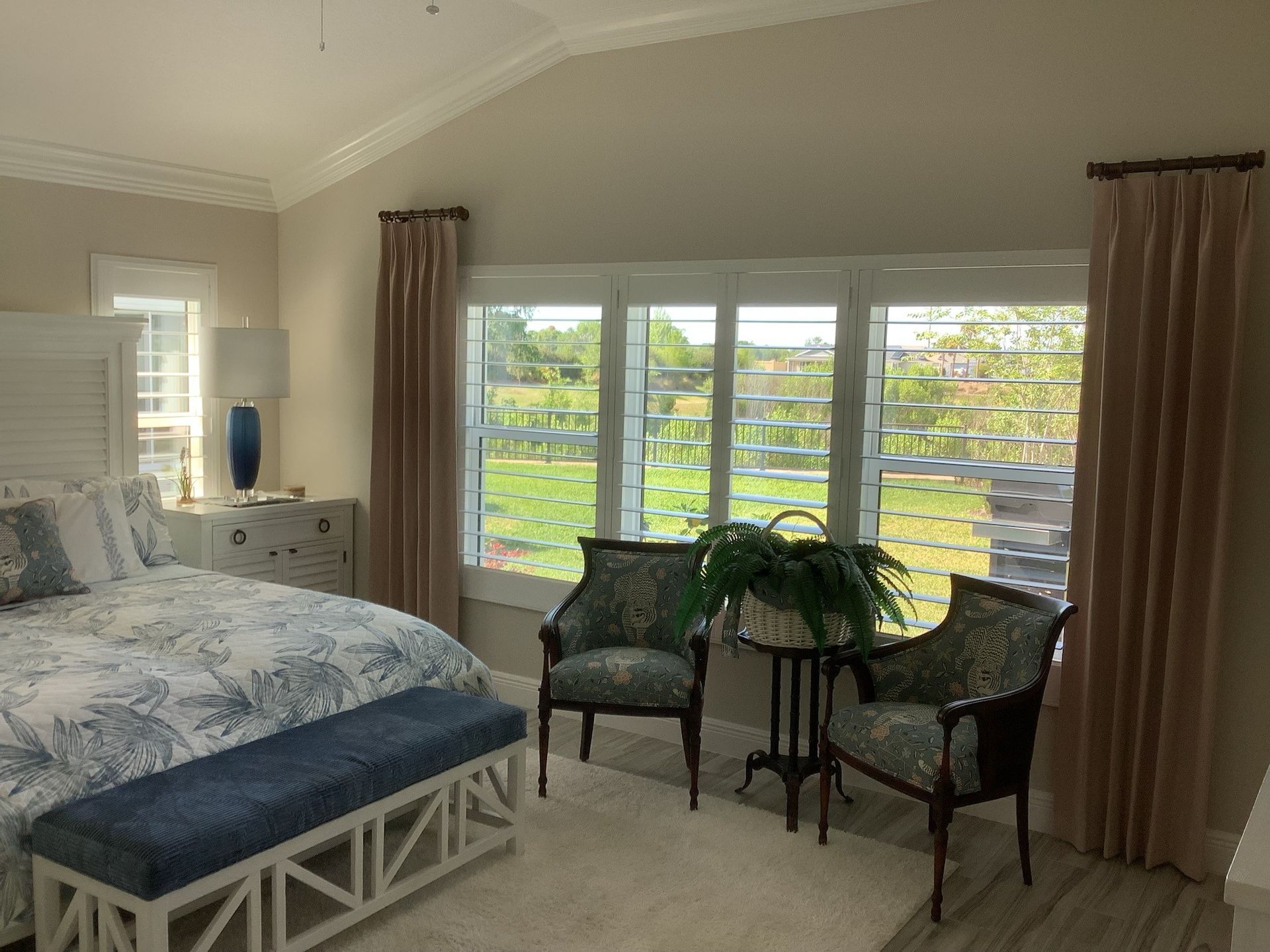 Bedroom with bed, two chairs, and curtains. Neutral walls, blue and white bedding, and outdoor view.