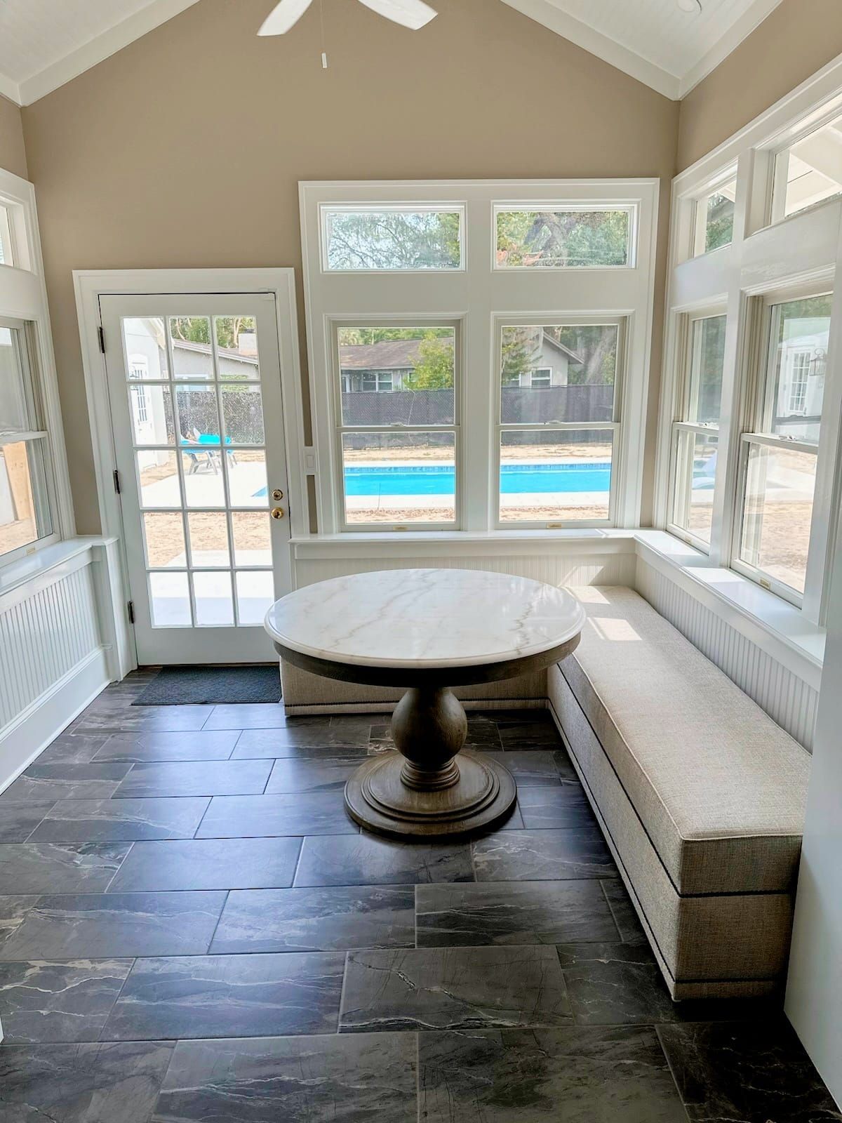 Sunroom with round table and built-in bench, facing a pool. Gray-tiled floor and large windows.
